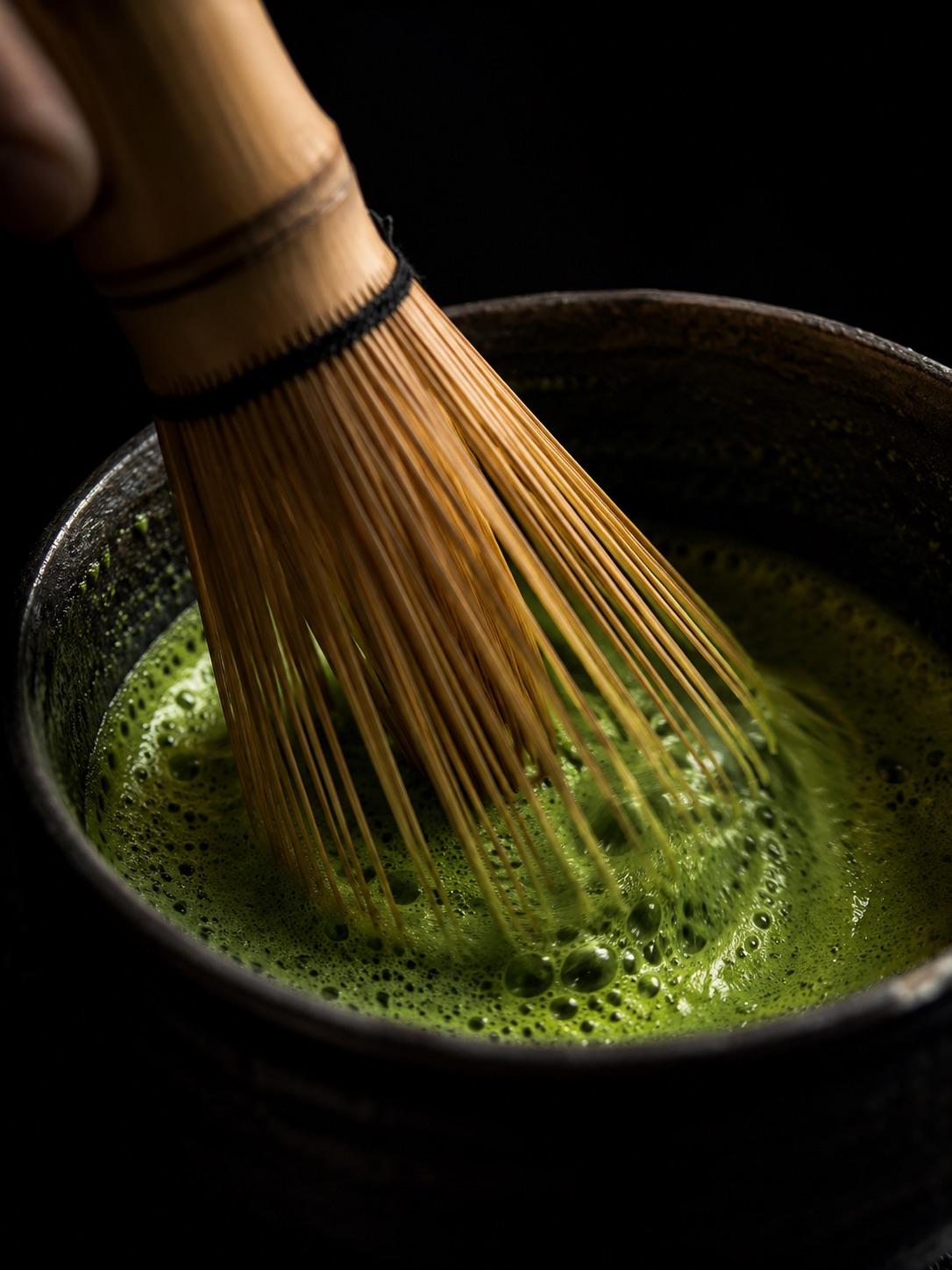 A bamboo chasen whisk creating bright green matcha foam in a dark bowl