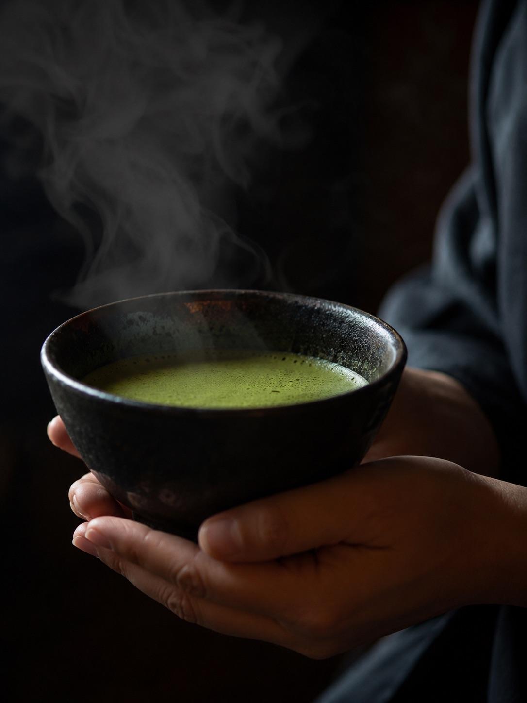 Hands holding a dark ceramic bowl with vibrant green matcha and soft steam
